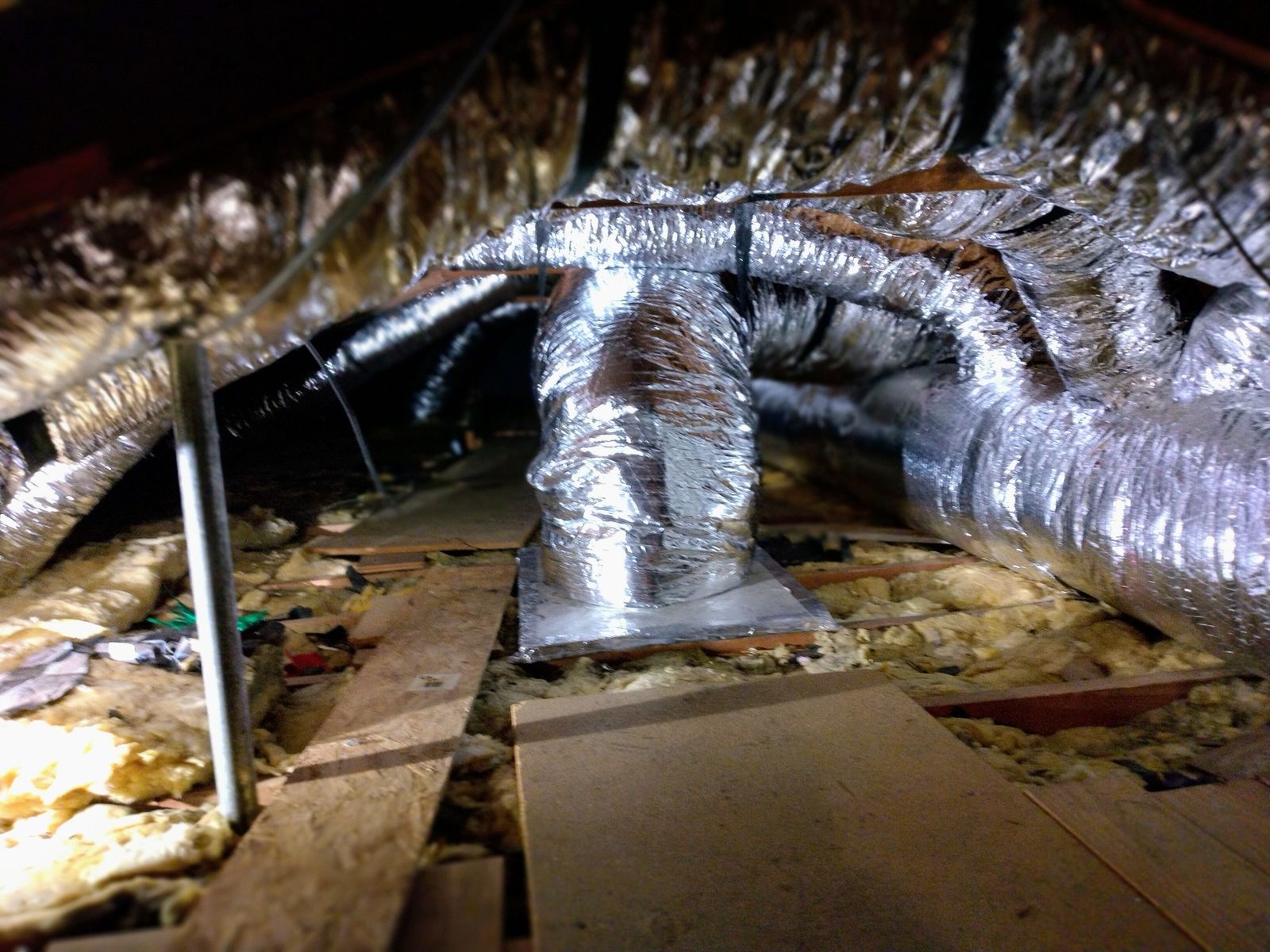 Attic with freshly installed shiny silver air ducts and exposed insulation. Wooden walk-boards form a path on the floor. Dimly lit, despite a bright work light.